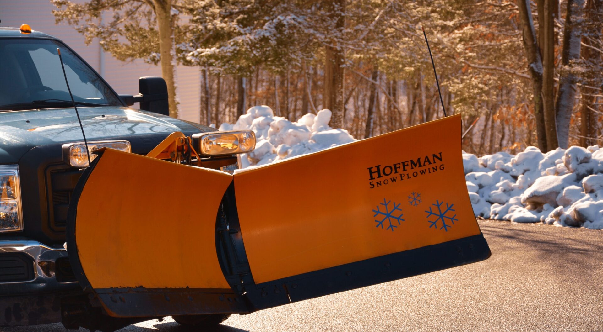 A snow plow truck with an orange and black blade reading "Hoffman Snow Plowing" is on a road surrounded by snowy trees with a clear sky.