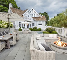 An elegant outdoor patio with wicker furniture, a fire pit, and a dining area against the backdrop of a luxurious white house with greenery.