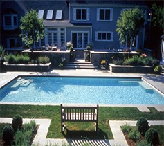 The image shows a backyard with a rectangular swimming pool, garden benches, manicured lawn, and a two-story house with outdoor seating and large windows.