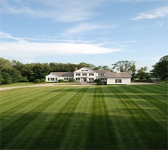 The image showcases a large, elegant two-story house with white siding amidst an expansive, manicured lawn with precise mowing patterns under a clear sky.