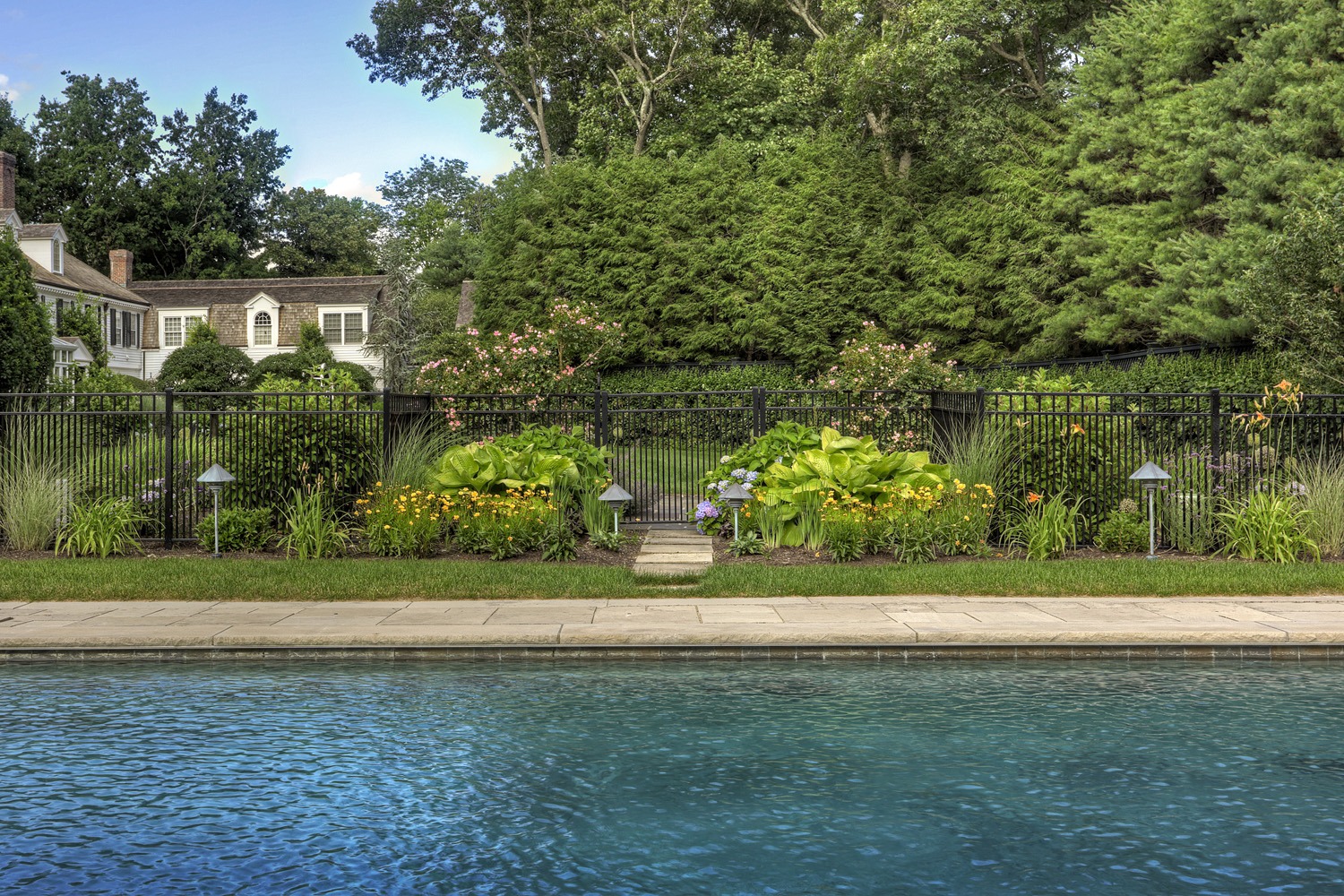An inviting outdoor swimming pool with crystal clear water bordered by a well-manicured lawn, vibrant flowering plants, and an elegant wrought-iron fence.
