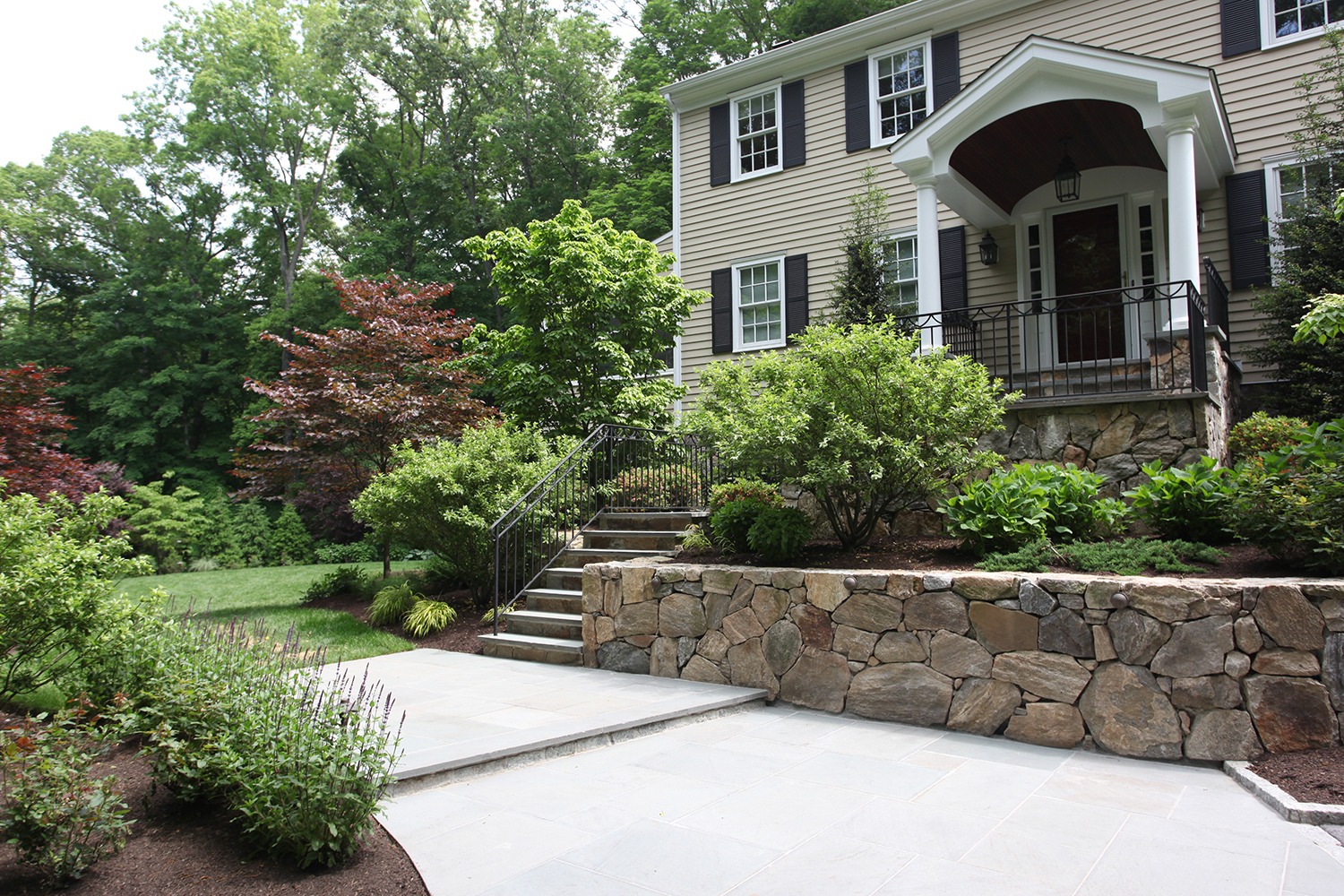 This image shows an elegant two-story house with a beige facade, white trim, and black shutters, surrounded by lush landscaping and stone work.