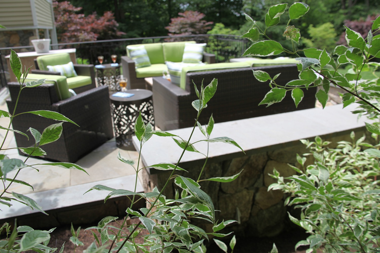 A patio with modern wicker furniture and green cushions. Plants foreground the setting. An ornate side table and wine glass are visible.