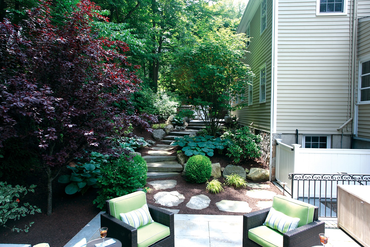 This image features a landscaped backyard with a stone pathway, green shrubbery, outdoor furniture, and a beige house with white trim.