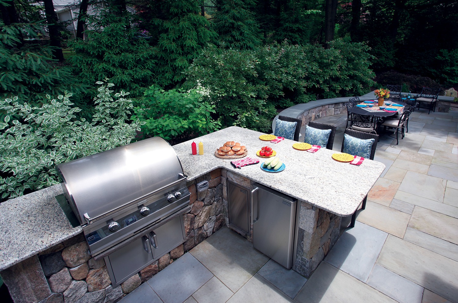 Outdoor kitchen with a built-in grill, stone countertops, and stainless-steel appliances. The dining area has a table set for a meal amid greenery.