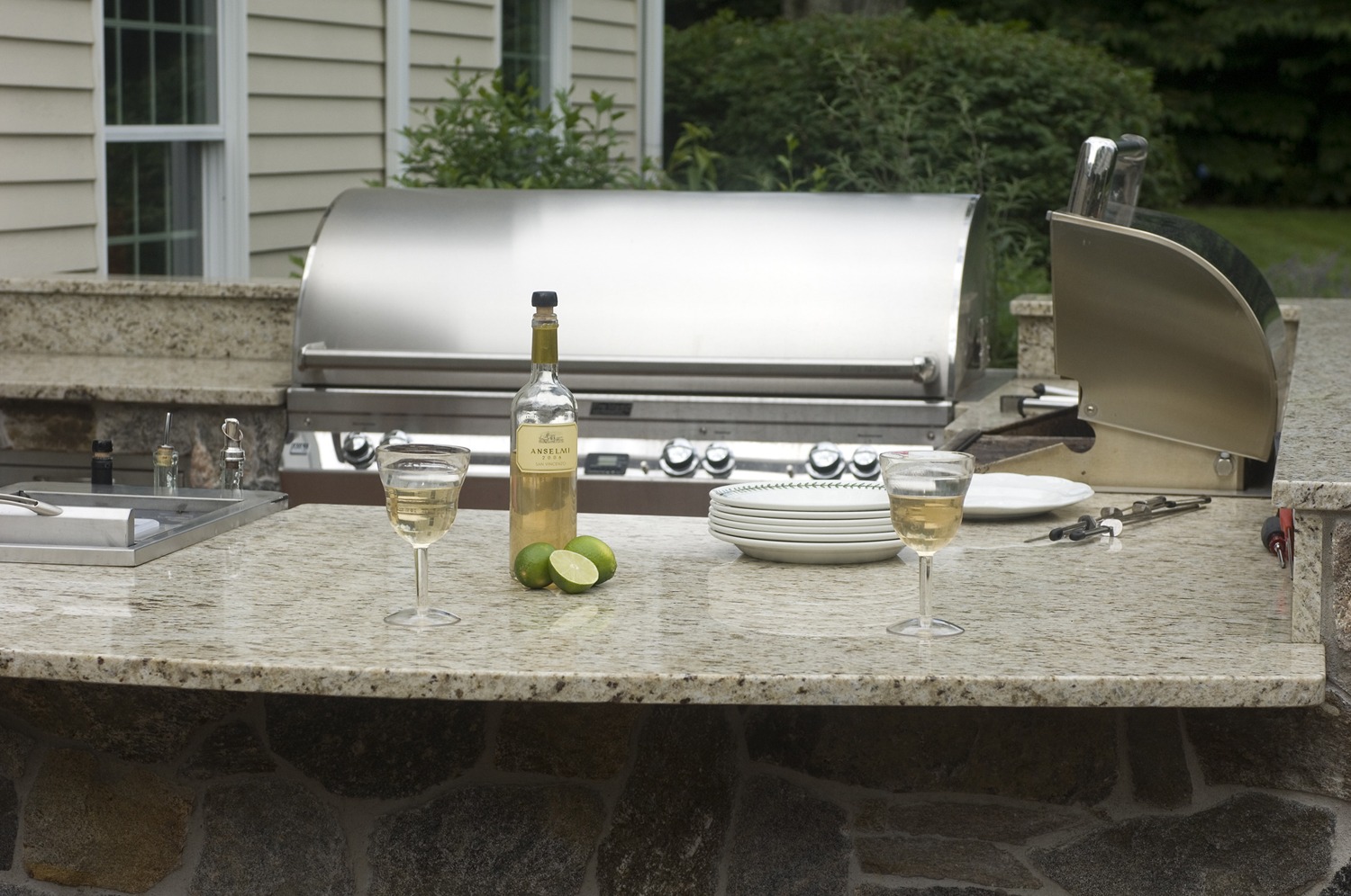 An outdoor kitchen setup showing a stainless steel grill, two glasses of white wine, a wine bottle, lime halves, and stacked plates on a granite countertop.