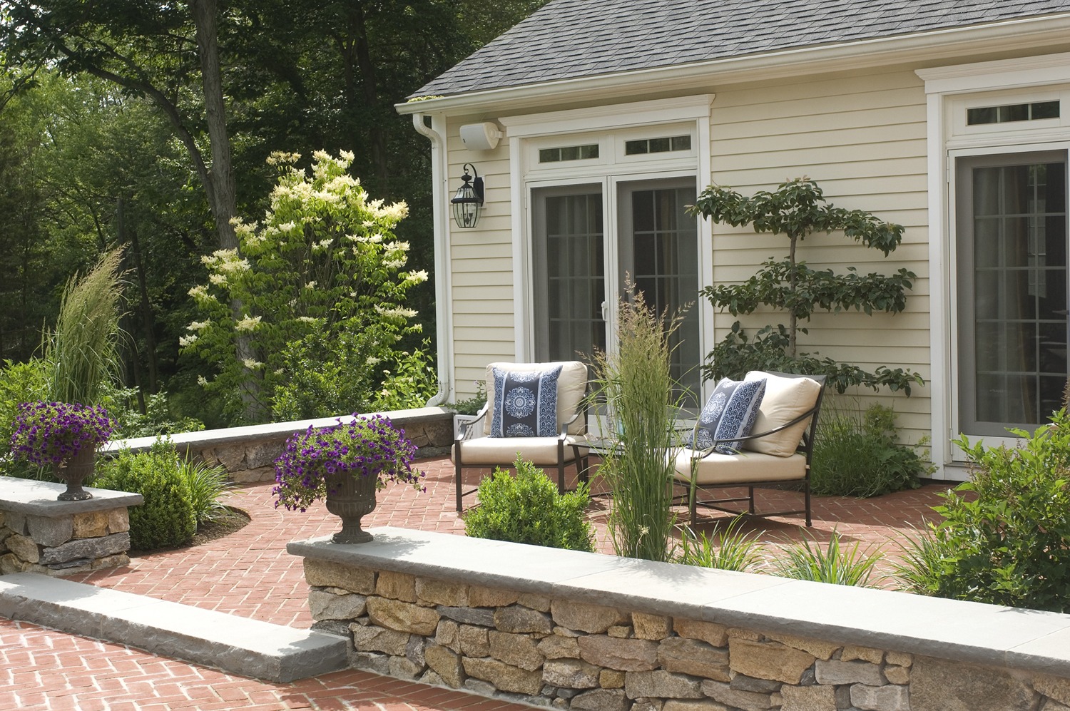 This is a serene patio area with a stone wall, comfortable seating, lush plants, and flower pots, adjacent to a house with double doors.