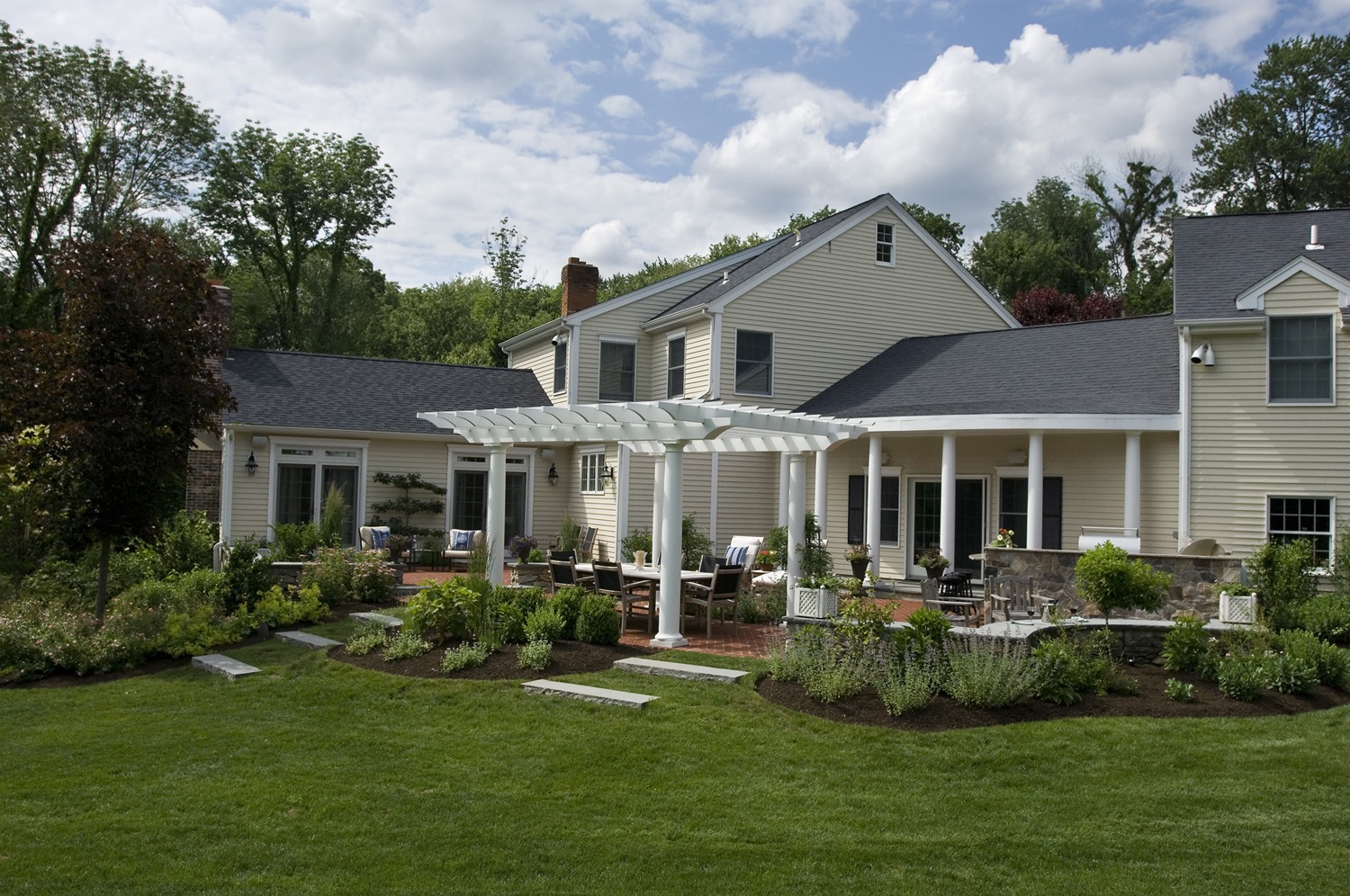 A traditional two-story house with beige siding features a covered patio area, manicured lawn, landscaped garden beds, and a pergola structure.