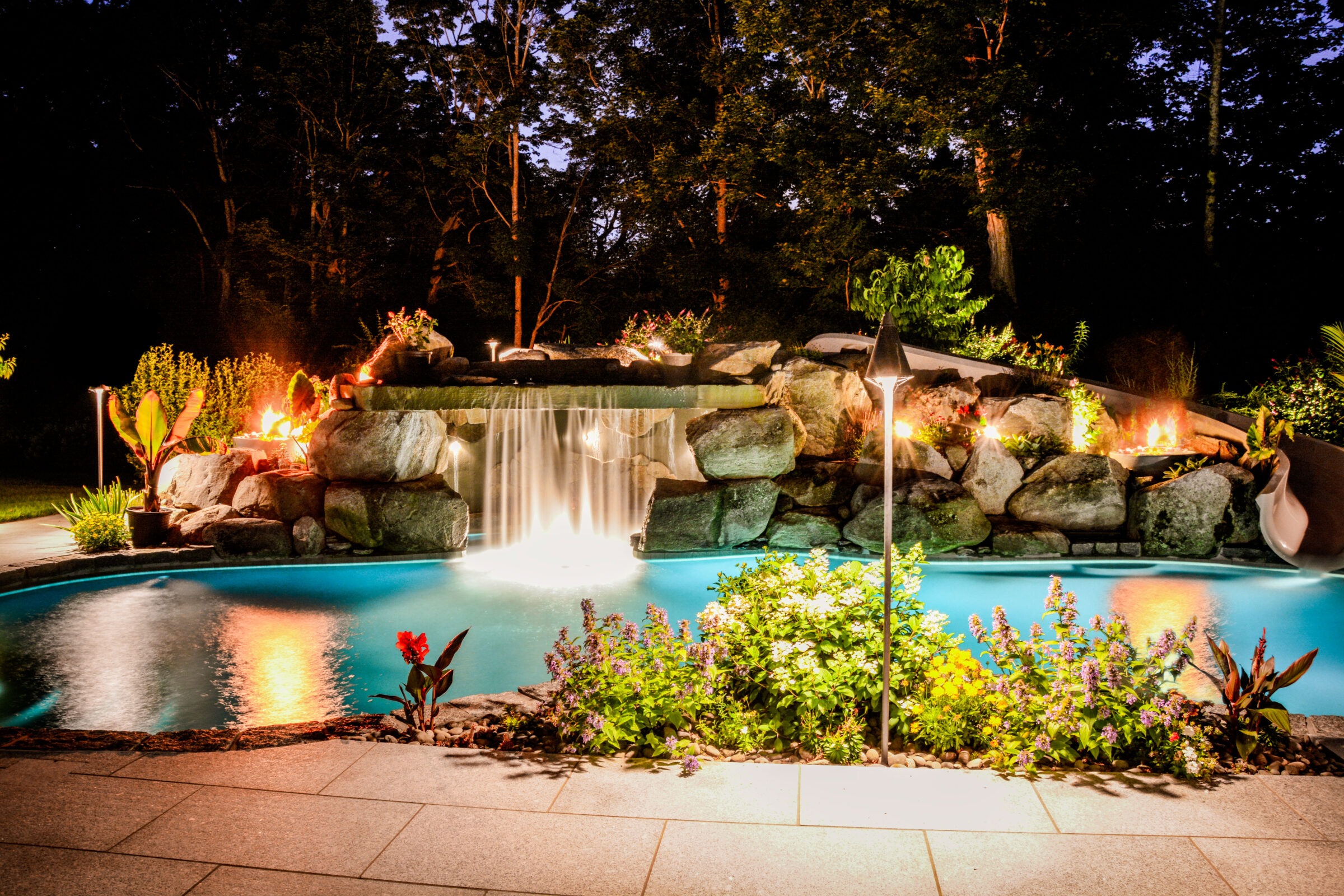 A luxuriously lit backyard pool at dusk, with a waterfall cascading over rocks, surrounded by lush plants, and tiki torches enhancing the ambience.