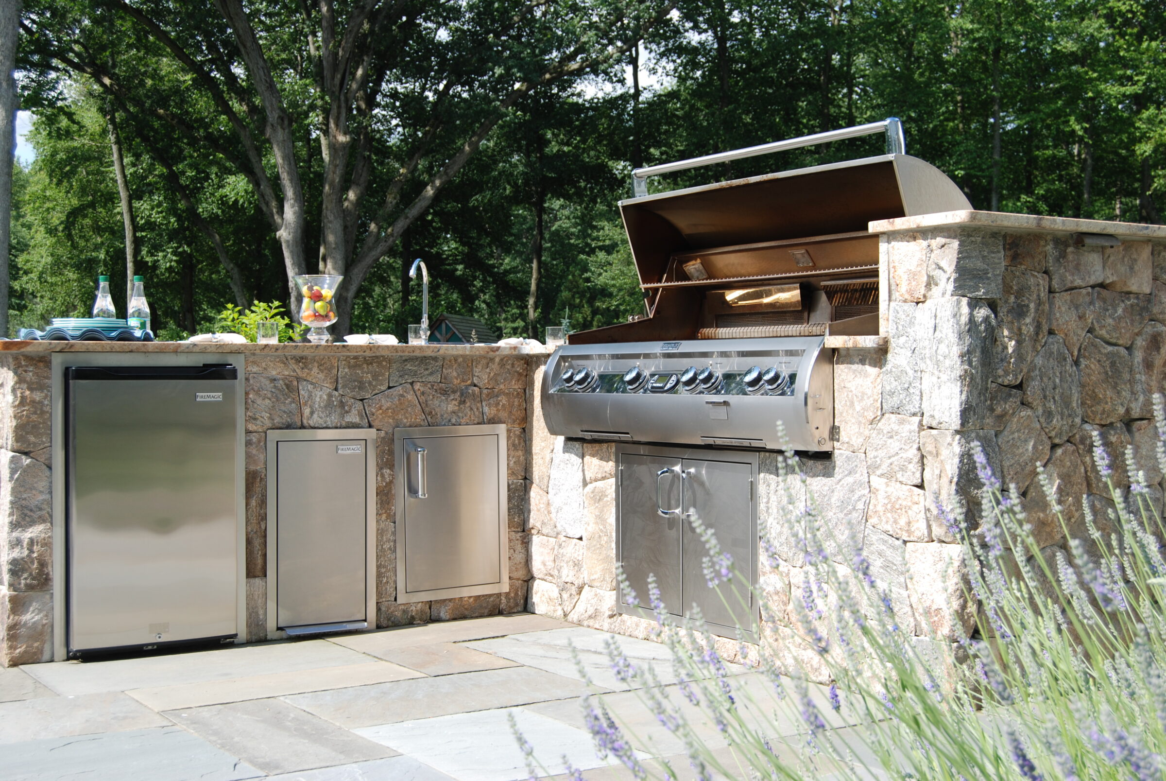 This image shows an outdoor kitchen with a built-in grill, refrigerator, and storage cabinets, set against a backdrop of trees and a clear sky.