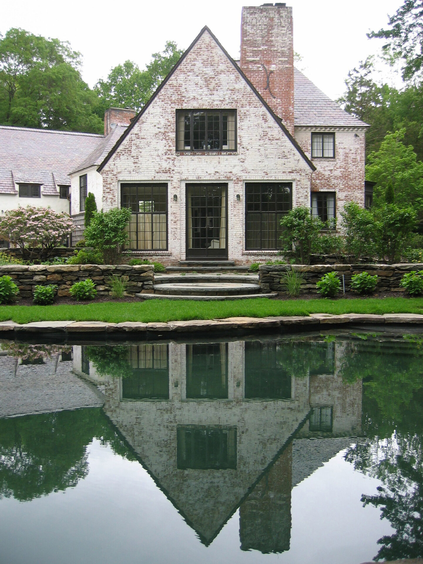 A traditional white brick house with a steep roof, reflected perfectly in a calm pond in front, surrounded by greenery and a stone wall border.