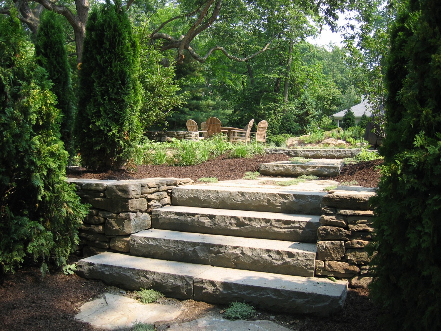 A landscaped garden with stone steps flanked by green shrubs. Wooden Adirondack chairs visible in the background under a tree canopy.