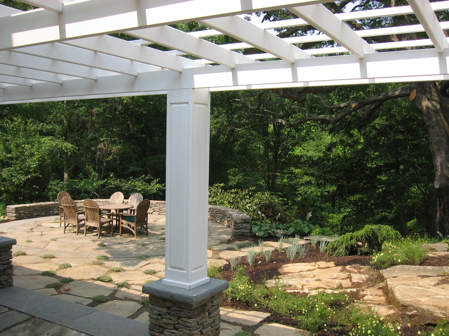 An outdoor patio with a pergola, wooden dining furniture, a stone pathway, landscaped garden beds, and dense trees in the background in daylight.
