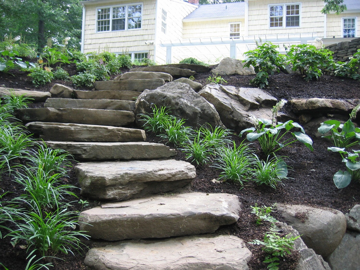 A natural stone staircase meanders through lush landscaping, leading towards a house with white siding and windows under a cloudy sky.