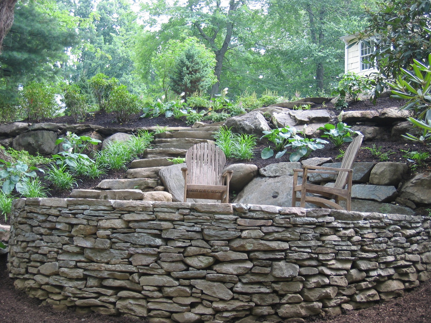 An outdoor stone wall and steps lead to a lush garden. Two wooden chairs invite relaxation amidst green trees and shrubs in a tranquil setting.