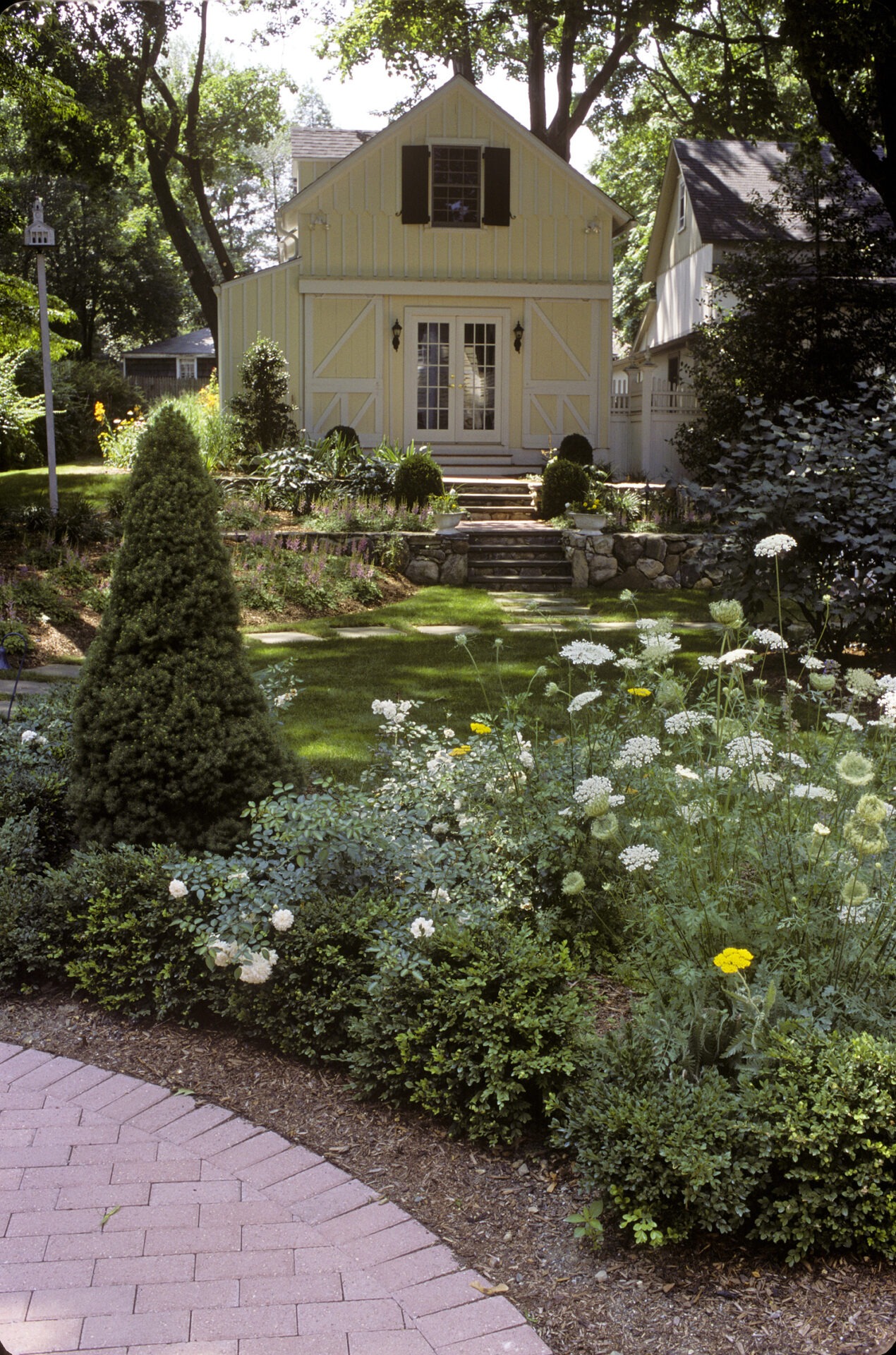 A quaint yellow house with white trim stands behind a lush garden with various plants and flowers, alongside a brick pathway, in a serene setting.