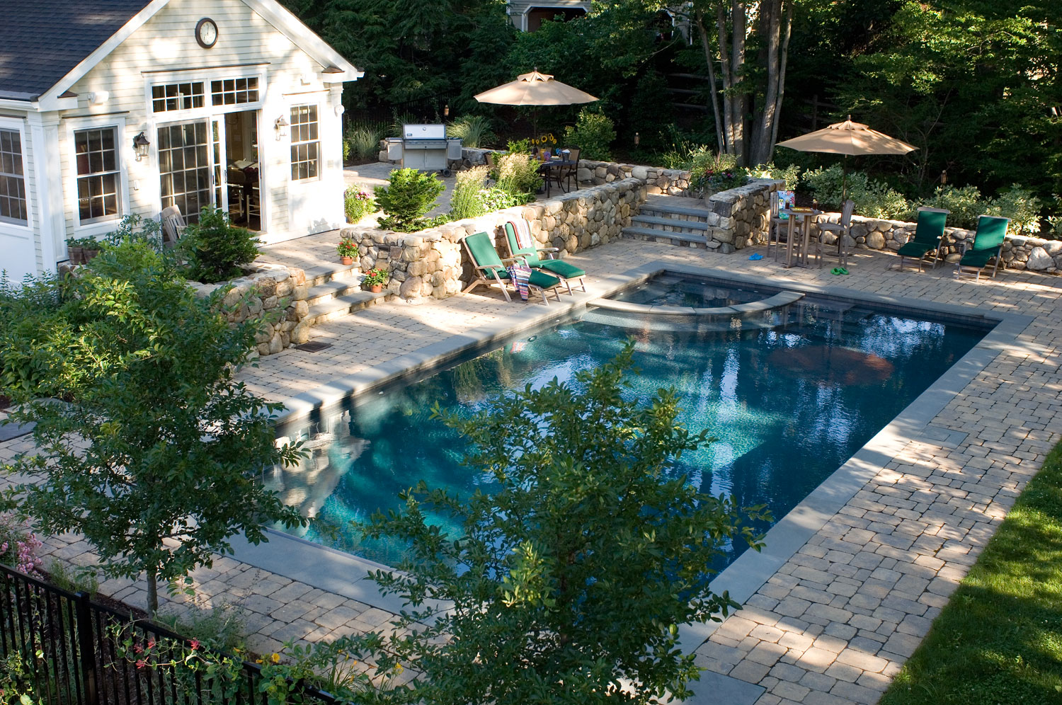 A serene backyard setting features an inviting rectangular swimming pool, stone patio, lush greenery, lounge chairs, and a quaint outbuilding.