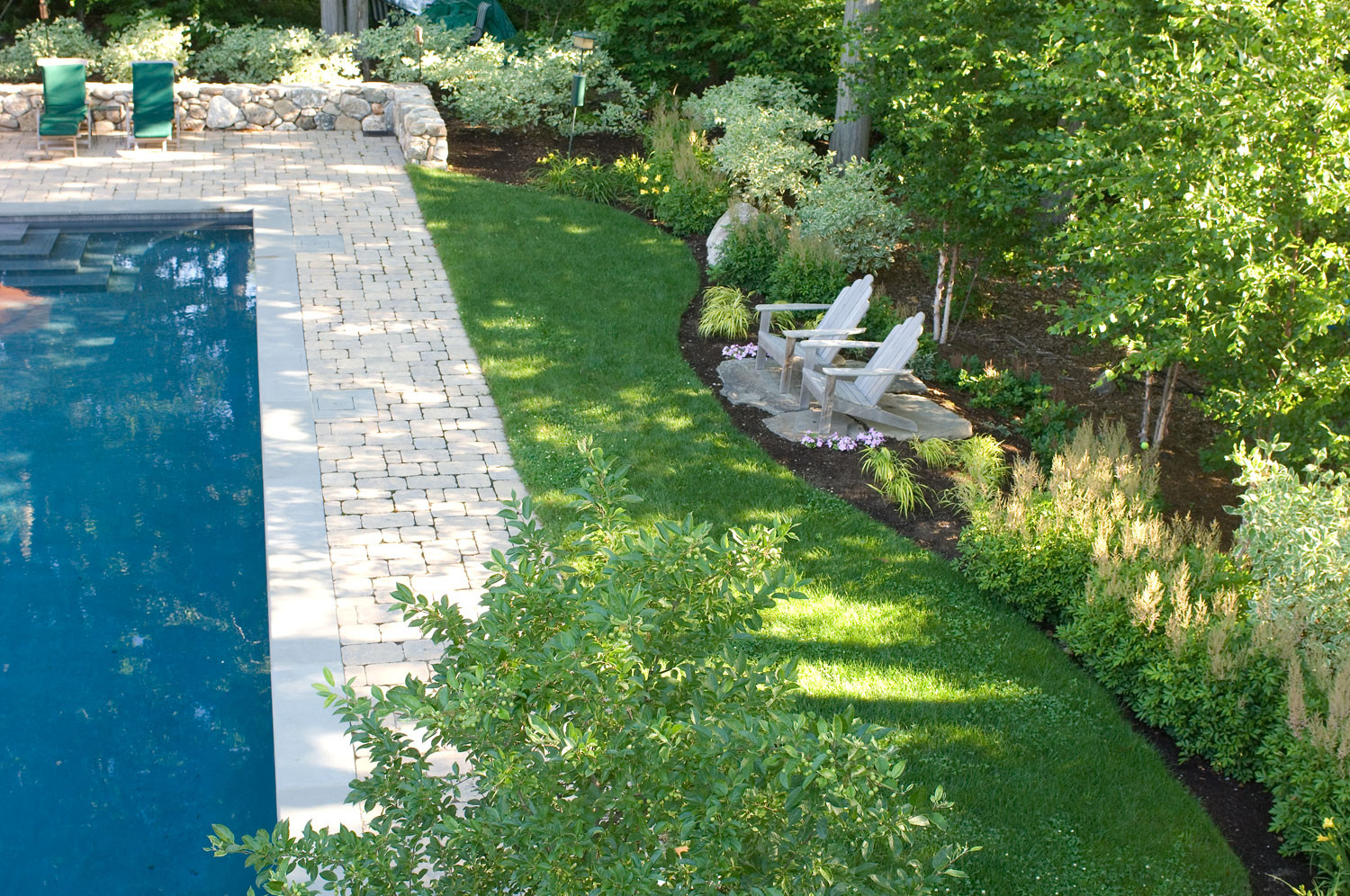 An outdoor scene with a clear blue swimming pool, stone patio, two Adirondack chairs, well-manicured grass, and lush garden beds under sunlight.