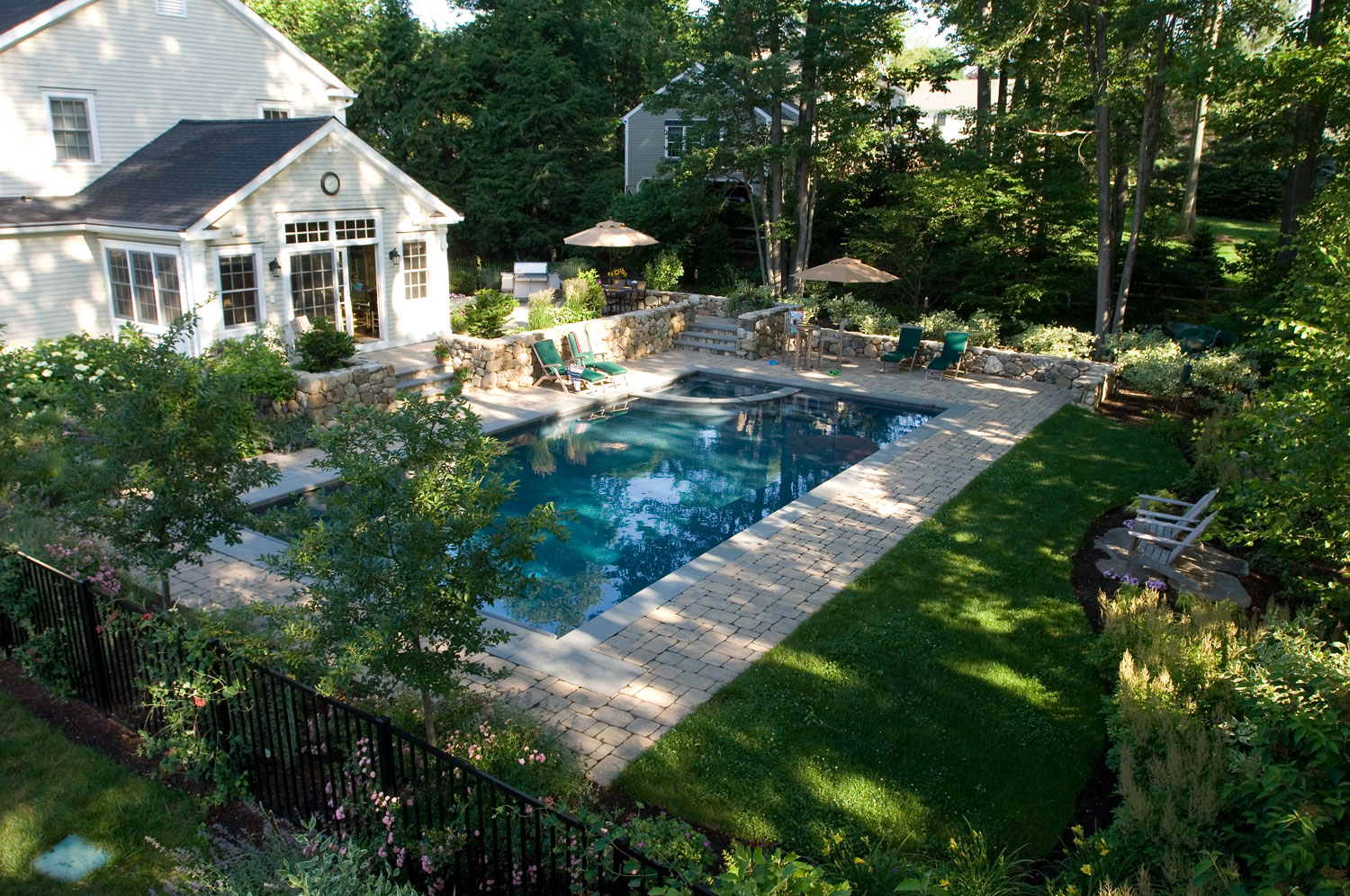 This image shows a well-landscaped backyard with an in-ground swimming pool, stone patio, lush greenery, outdoor furniture, and a two-story house.