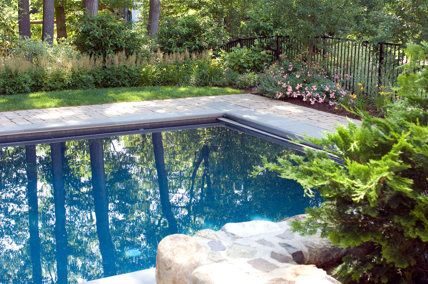A serene backyard setting featuring a covered swimming pool, landscaped garden, a stone pathway, lush greenery, and a decorative wrought-iron fence.