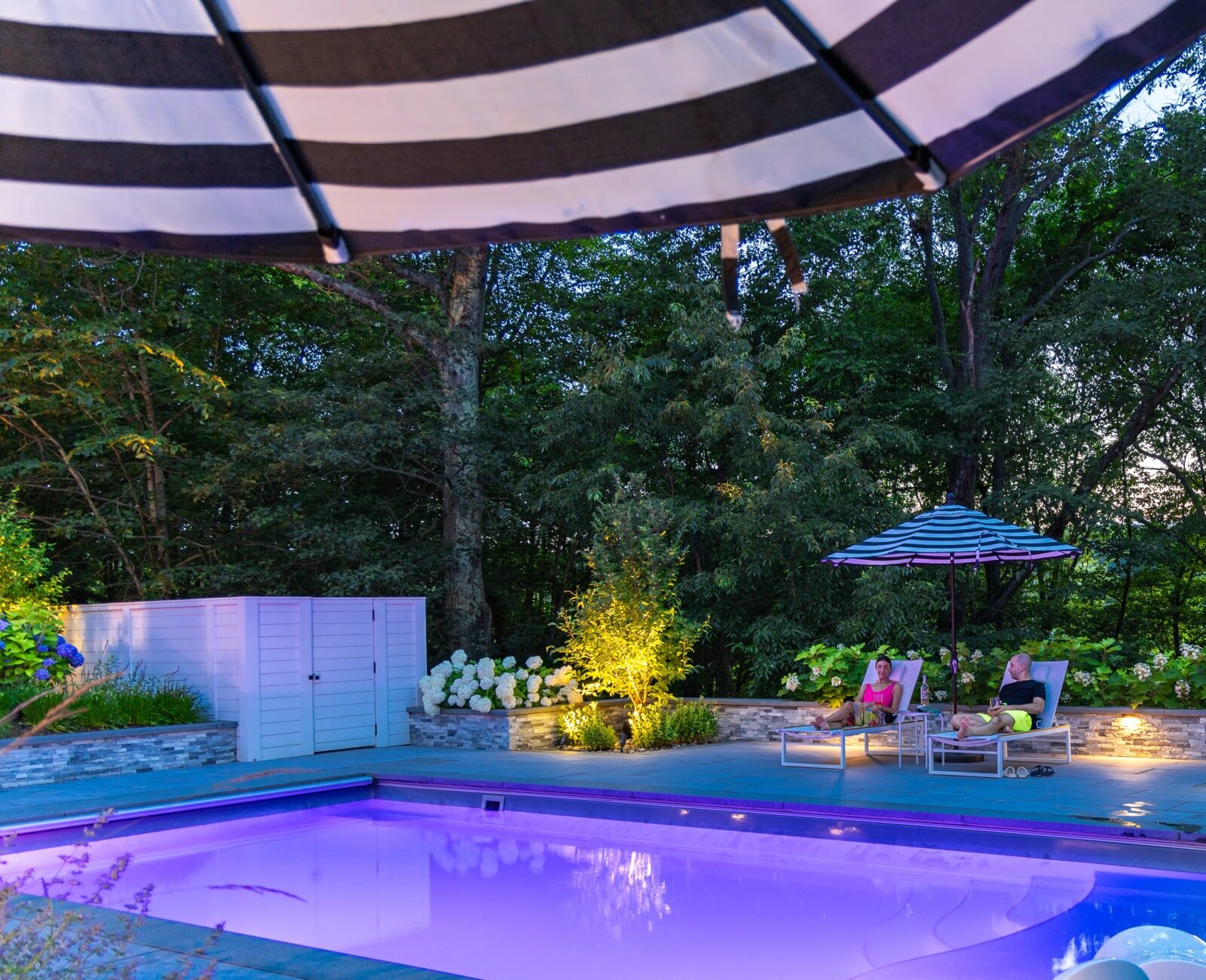 An outdoor pool area at dusk with illuminated landscaping, two people lounging under a striped umbrella, and a waterslide leading into the pool.