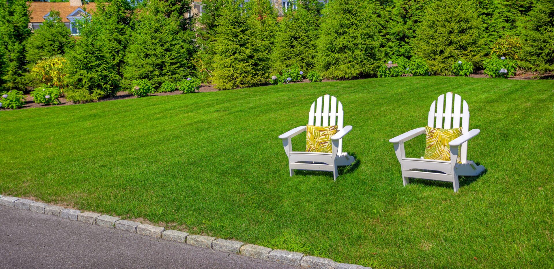 Two white Adirondack chairs with yellow-patterned pillows are placed on a neatly trimmed green lawn, with lush greenery and a house in the background.