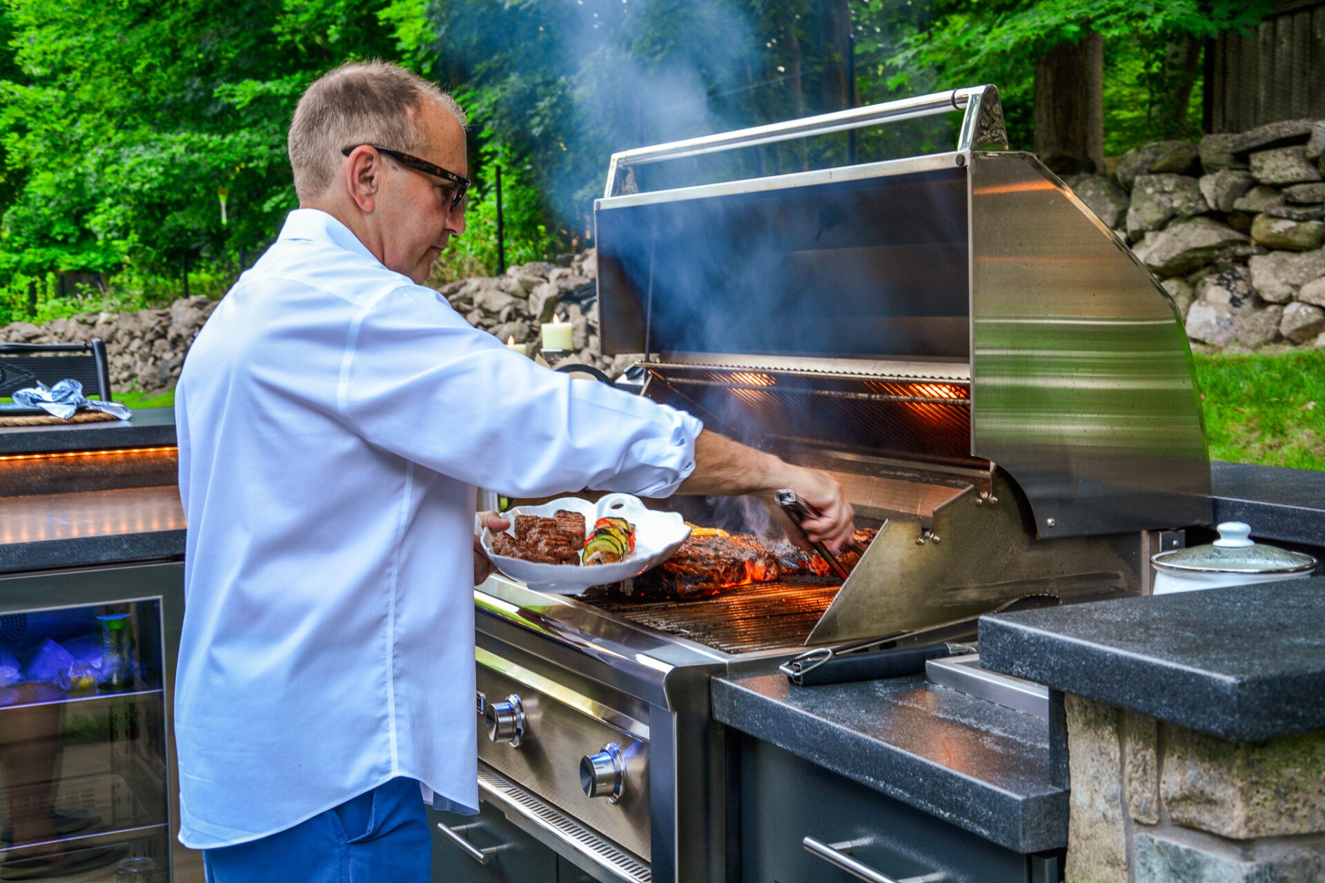 A person in a white shirt is grilling meat on an outdoor barbecue. The grill is smoking, and a platter is held in one hand.