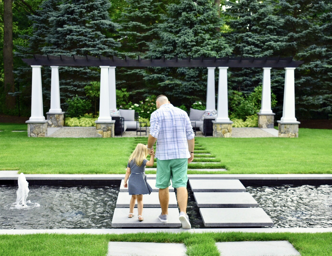 A person is walking with a child across stepping stones over a water feature in a lush garden with white columns and a pergola in the background.