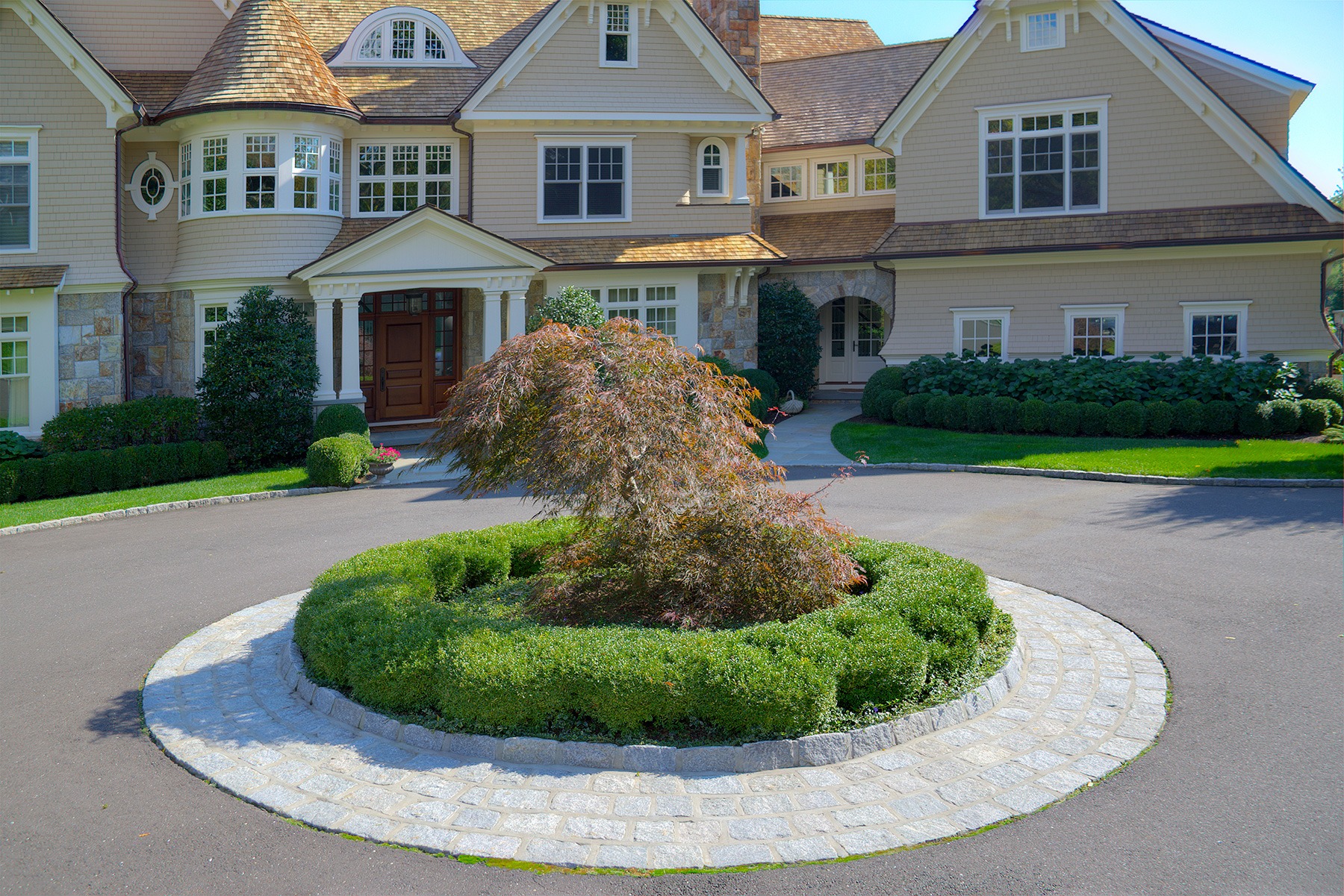 A large, elegant house with beige siding, white trim, a landscaped yard, circular driveway, and a central planter with a sculpted bush and tree.