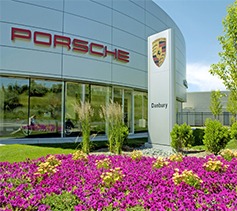 The image shows the exterior of a Porsche dealership with a well-maintained landscape featuring colorful flowers and green grass under a clear sky.