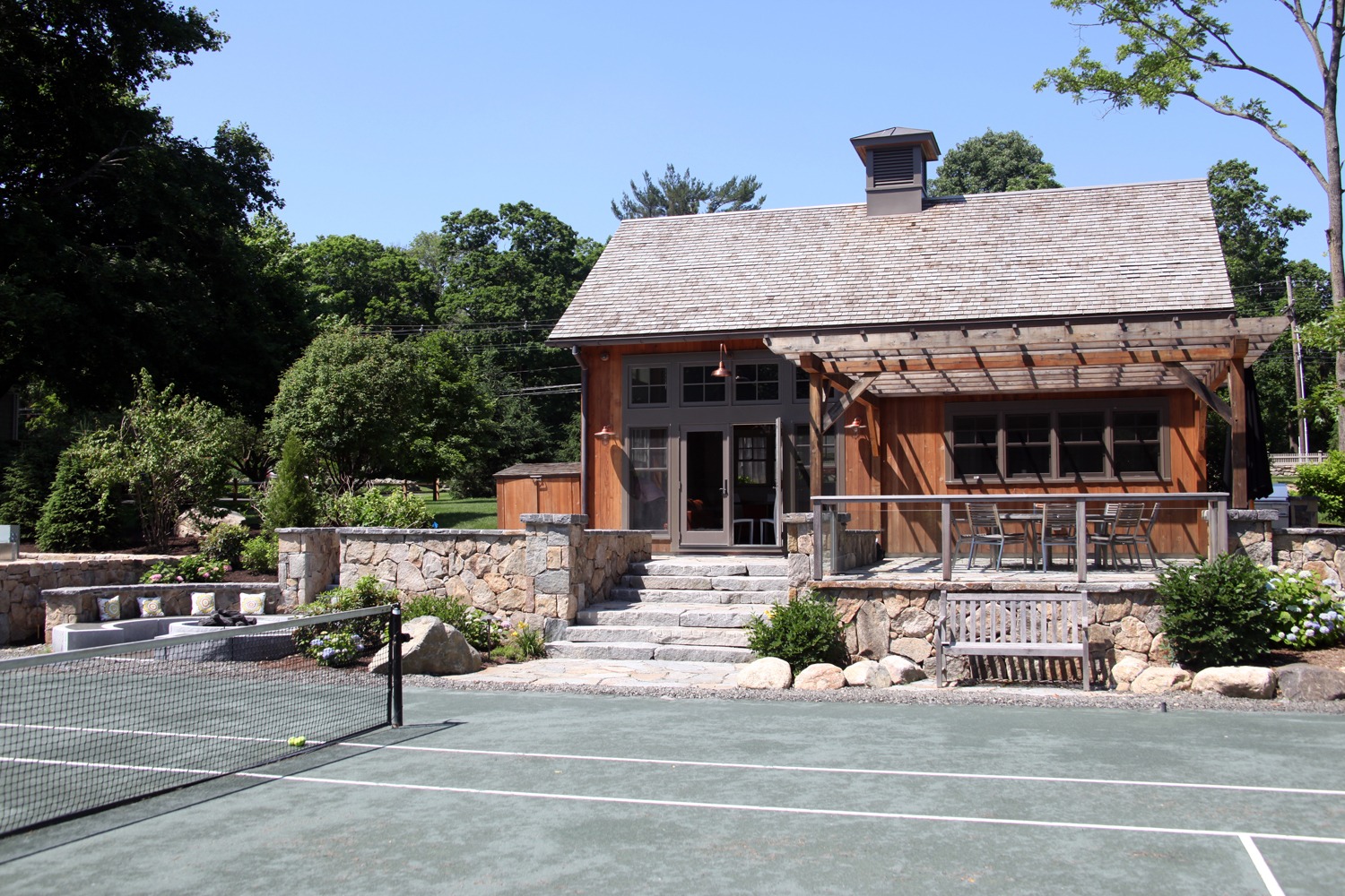 A rustic building with a cedar shake roof sits beside a tennis court, featuring stone stairs, landscaping, and a clear blue sky overhead.