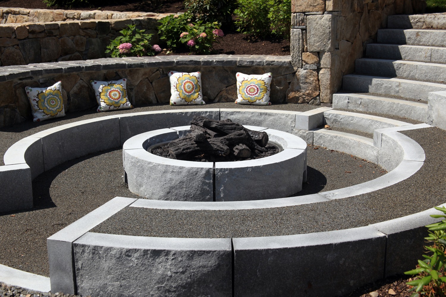 An outdoor stone fire pit sits at the center of curved bench seating with decorative pillows. Stone stairs lead up to an adjacent area.