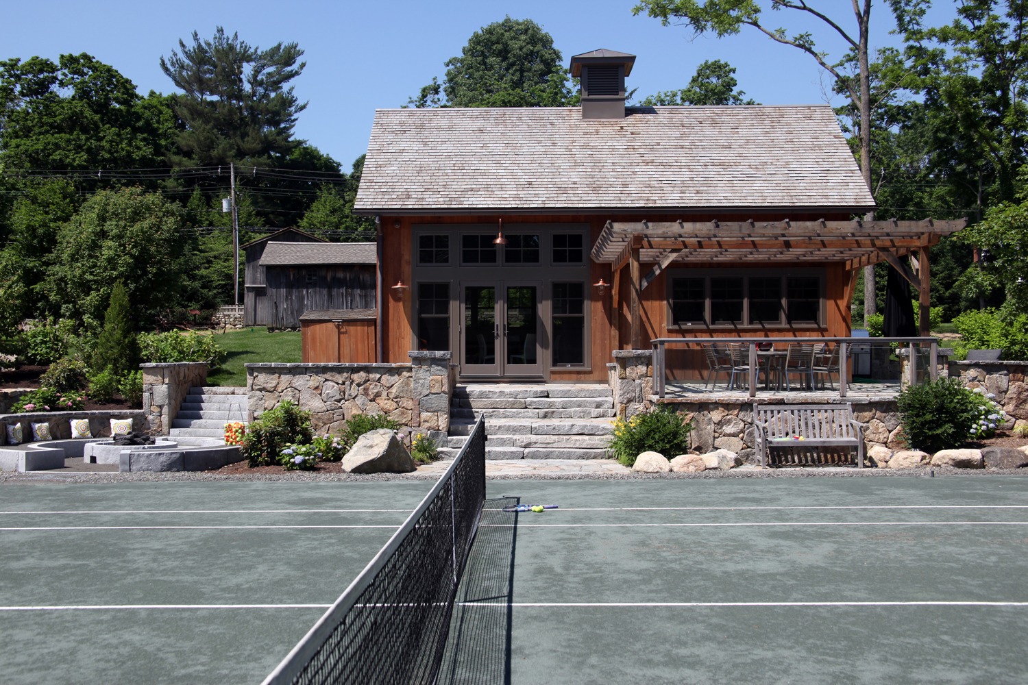 A tennis court with a wooden pavilion house, stone steps, benches, lush greenery, and a clear blue sky. Rackets rest on the ground near the net.