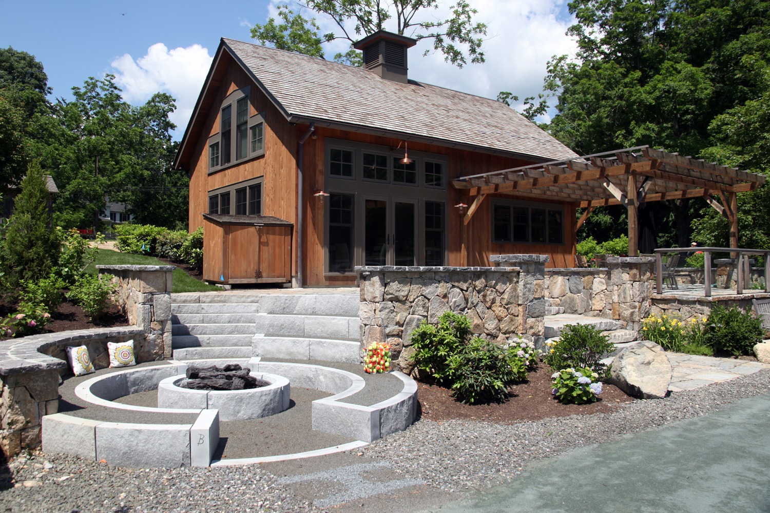 This image features a rustic wooden house with a stone foundation, a pergola, steps leading up to a porch, landscaping, and a circular fire pit.