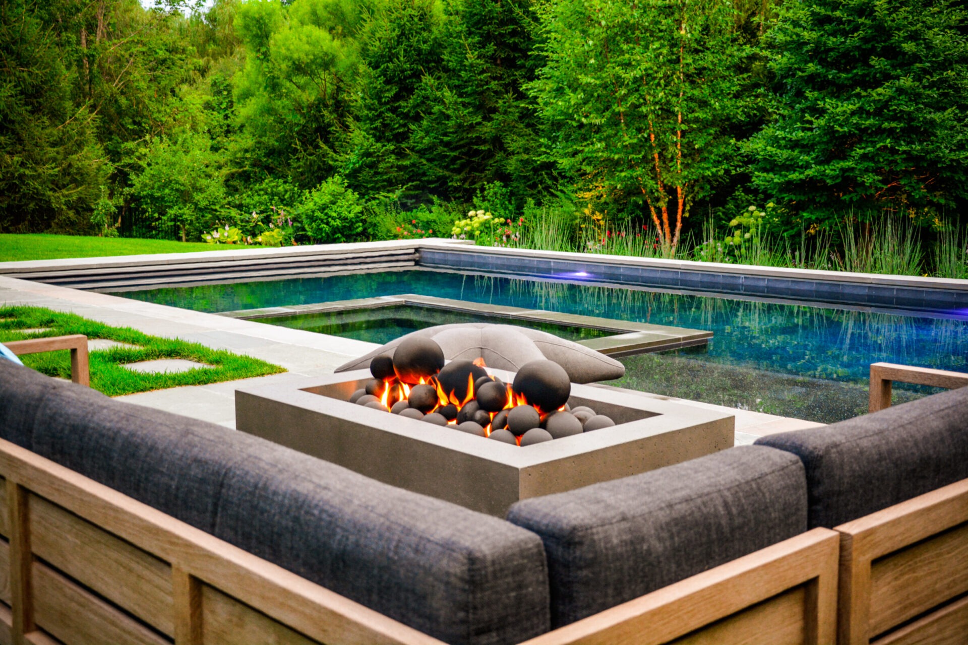 An outdoor fire pit with a flame lit, surrounded by sleek furniture overlooking a calm swimming pool set against a backdrop of lush greenery.