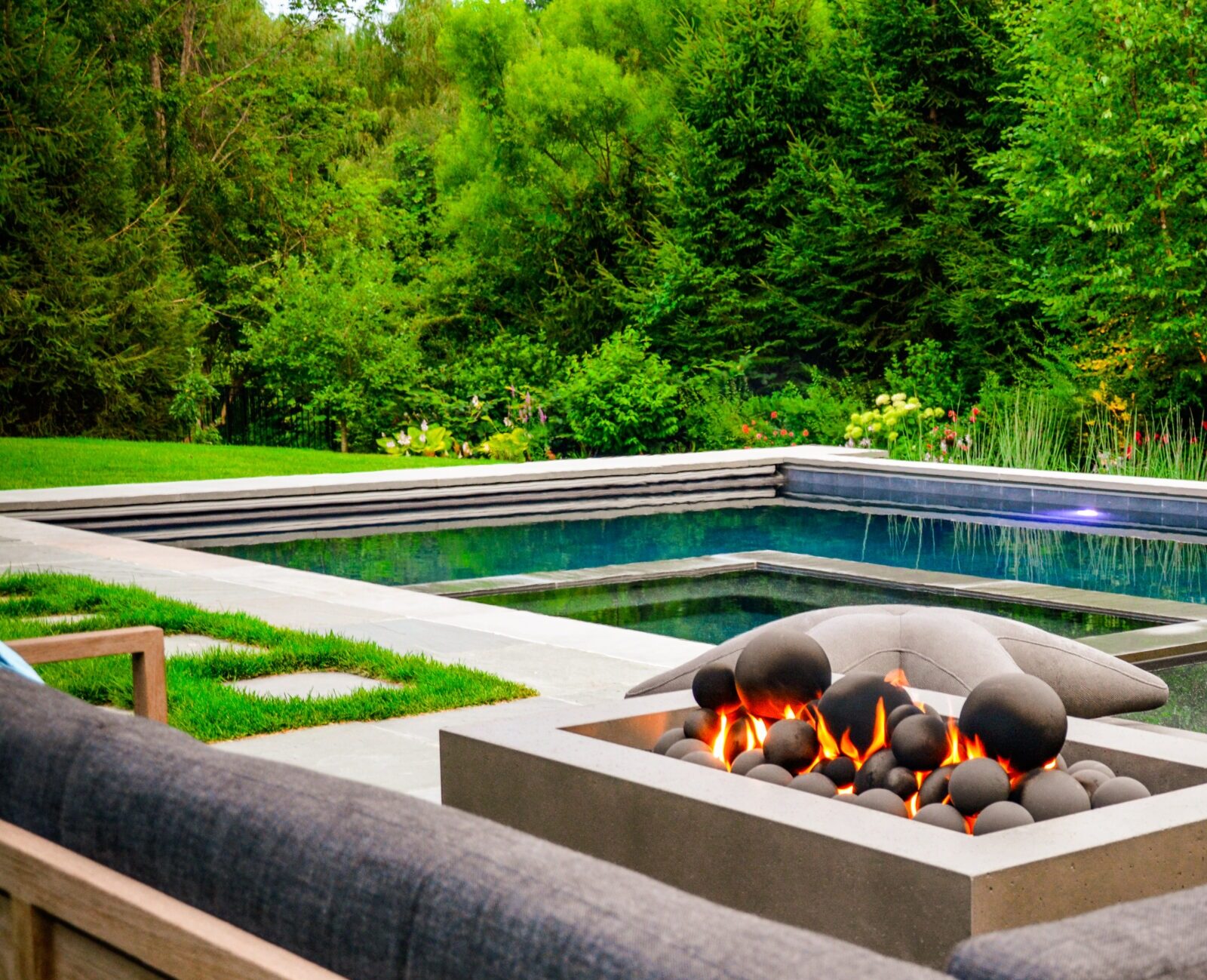 An outdoor fire pit with a flame lit, surrounded by sleek furniture overlooking a calm swimming pool set against a backdrop of lush greenery.
