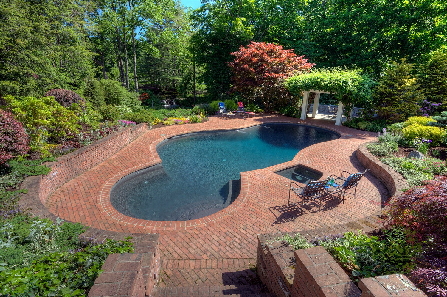 A kidney-shaped swimming pool surrounded by a brick patio and lush garden. Features include a covered pergola, outdoor furniture, and diverse plants.