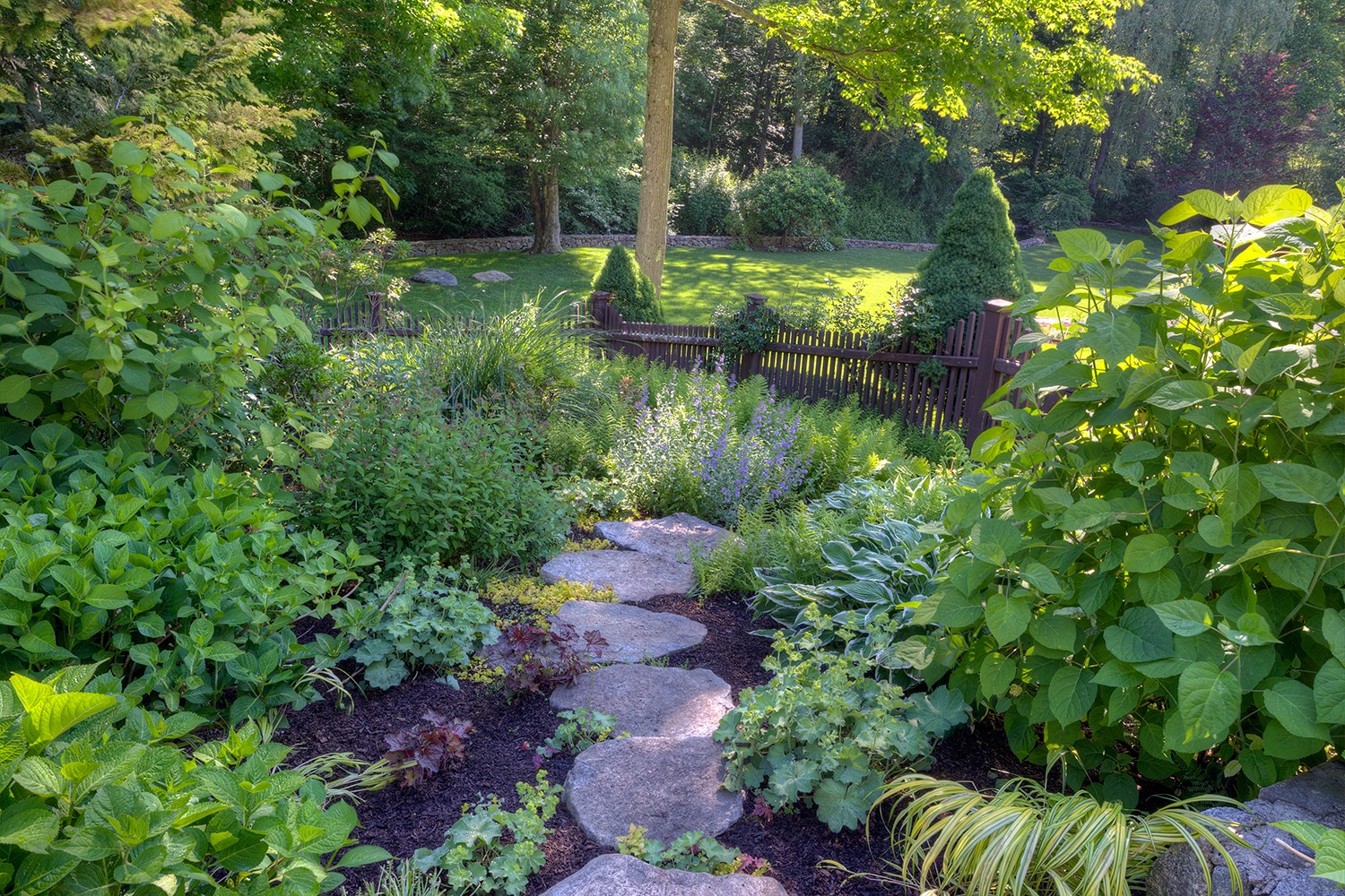 A serene garden with a stone path leading through lush vegetation to a neatly trimmed lawn, surrounded by a wooden fence, and shaded by trees.