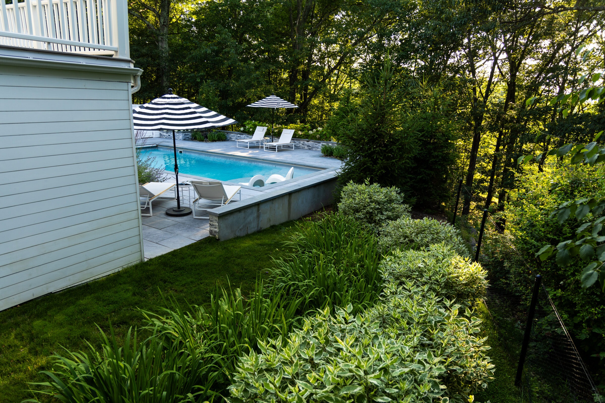 A serene backyard with a swimming pool, surrounded by lush greenery, featuring striped umbrellas, lounge chairs, and a well-manicured lawn.