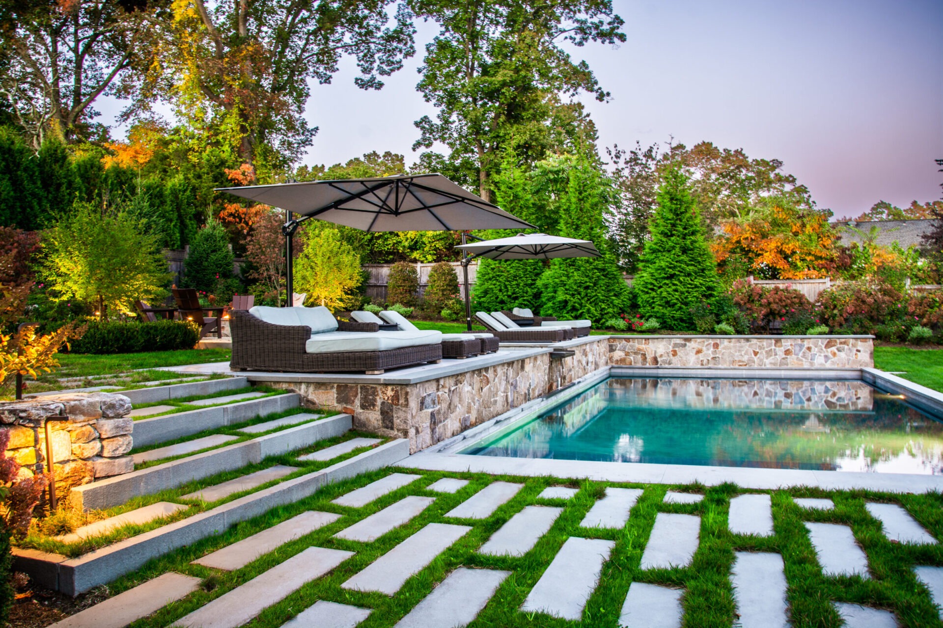 A luxurious backyard with a rectangular pool, stone pathways, outdoor furniture under an umbrella, lush greenery, and a well-manicured lawn at twilight.