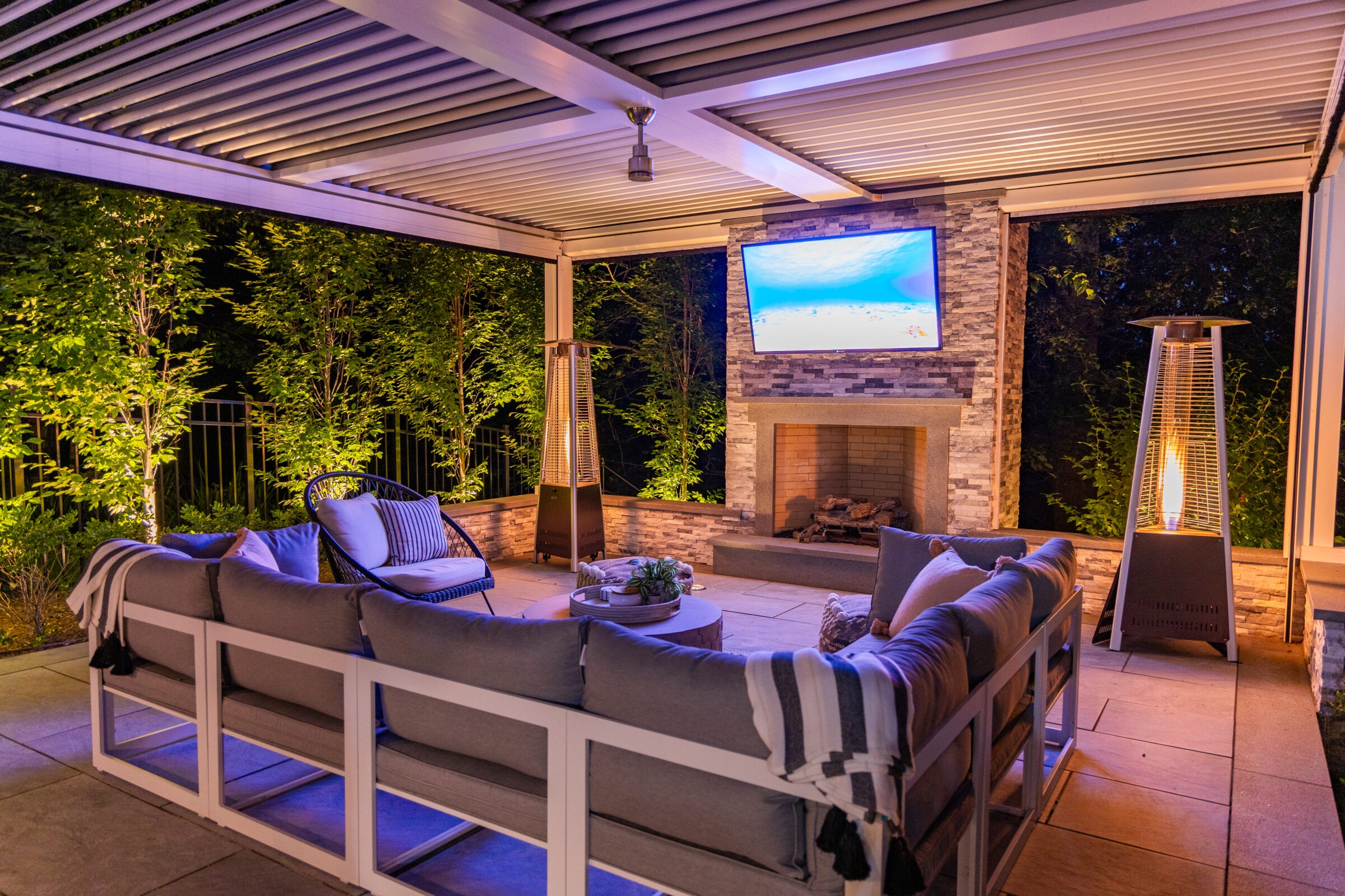 An elegant outdoor living space with comfortable seating, a fireplace, patio heater, and a television mounted above the fireplace for entertainment.