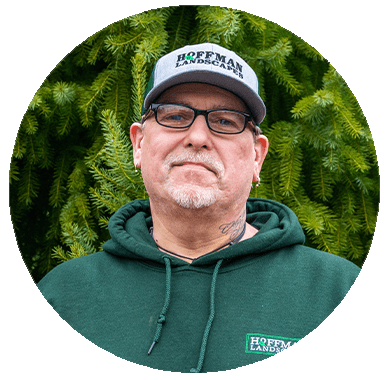 This is an image of a person wearing glasses, a cap, and a green hoodie with an embroidered logo. They have a tattoo on their neck.