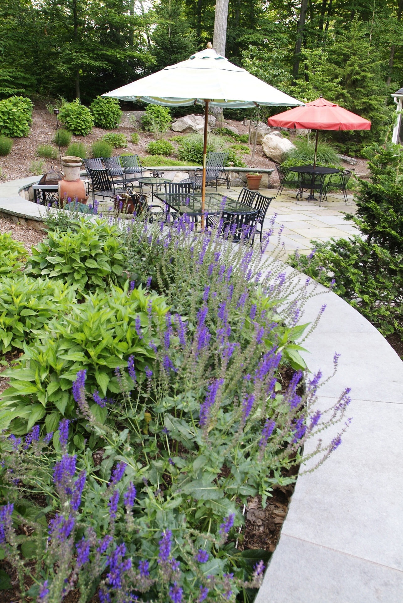 An outdoor patio area with purple flowers in the foreground, surrounded by greenery, featuring a seating area with umbrellas and metal furniture.