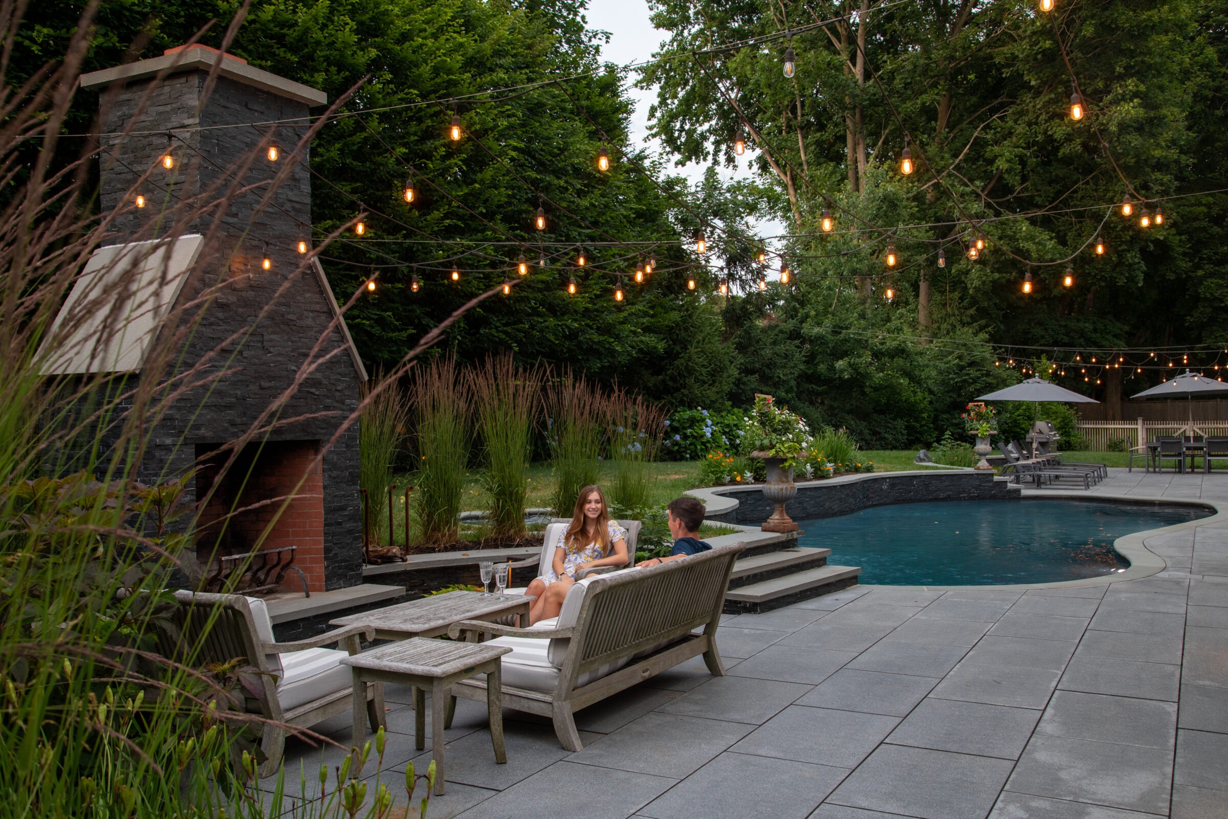 An outdoor setting with two people seated, a swimming pool, string lights, greenery, patio furniture, and an evening sky. It appears serene and leisurely.
