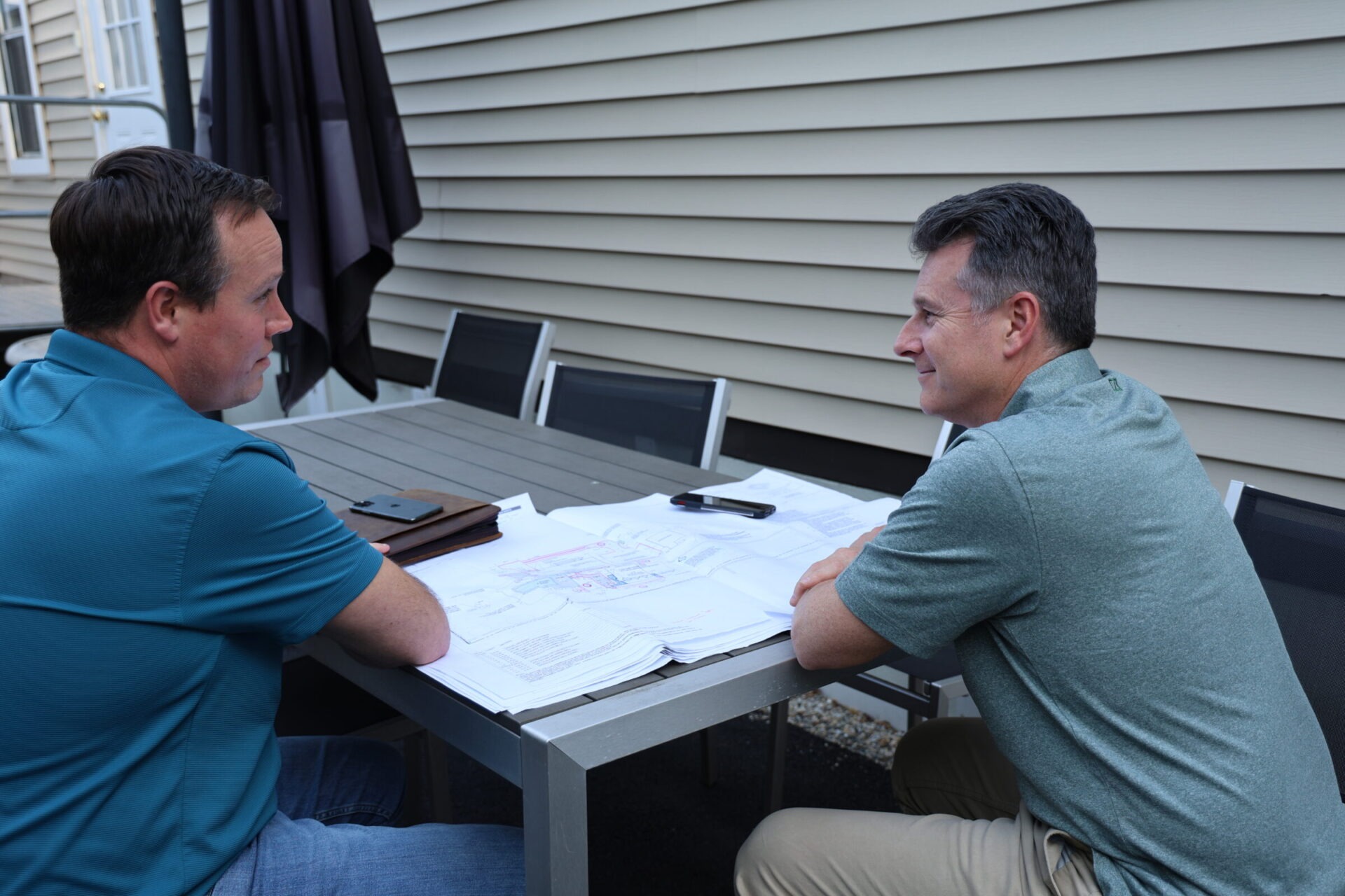 Two people sit at an outdoor table reviewing plans or documents, engaged in conversation, with a house facade and a patio umbrella in the background.