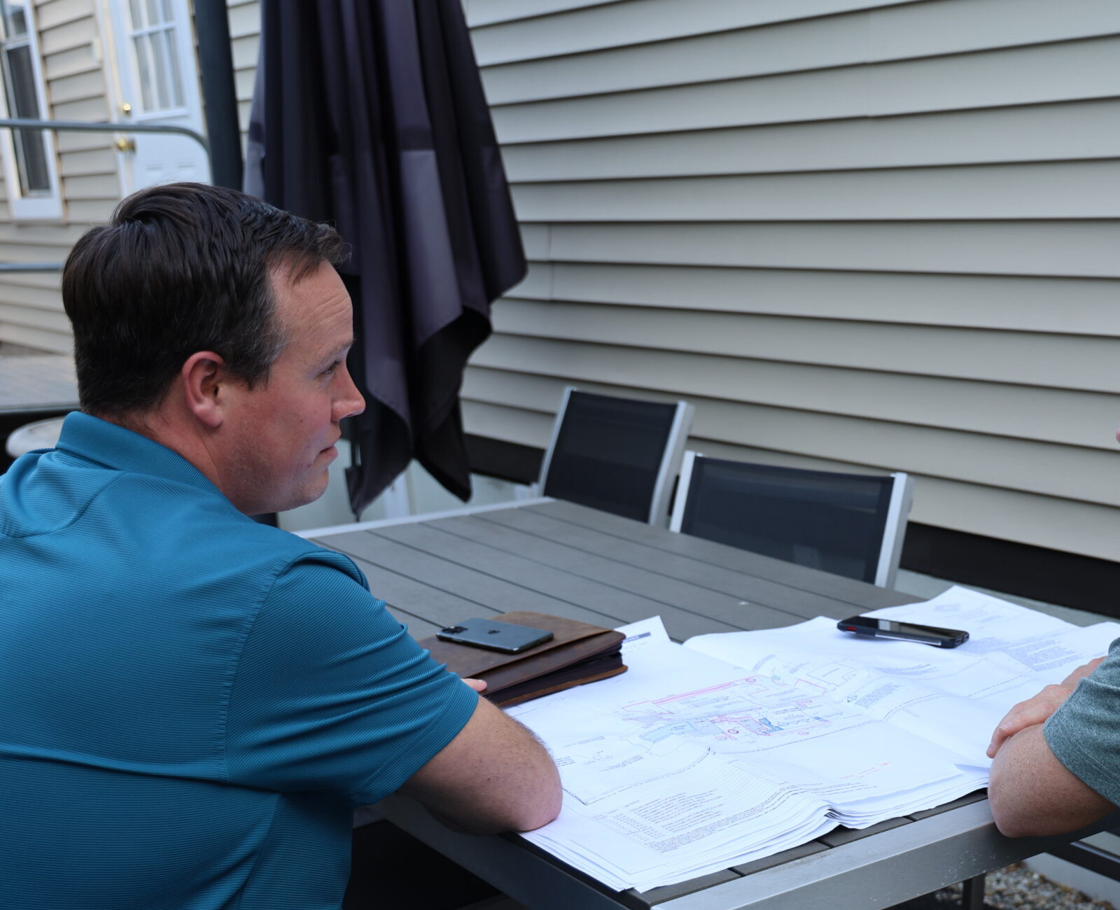 Two people sit at an outdoor table reviewing plans or documents, engaged in conversation, with a house facade and a patio umbrella in the background.
