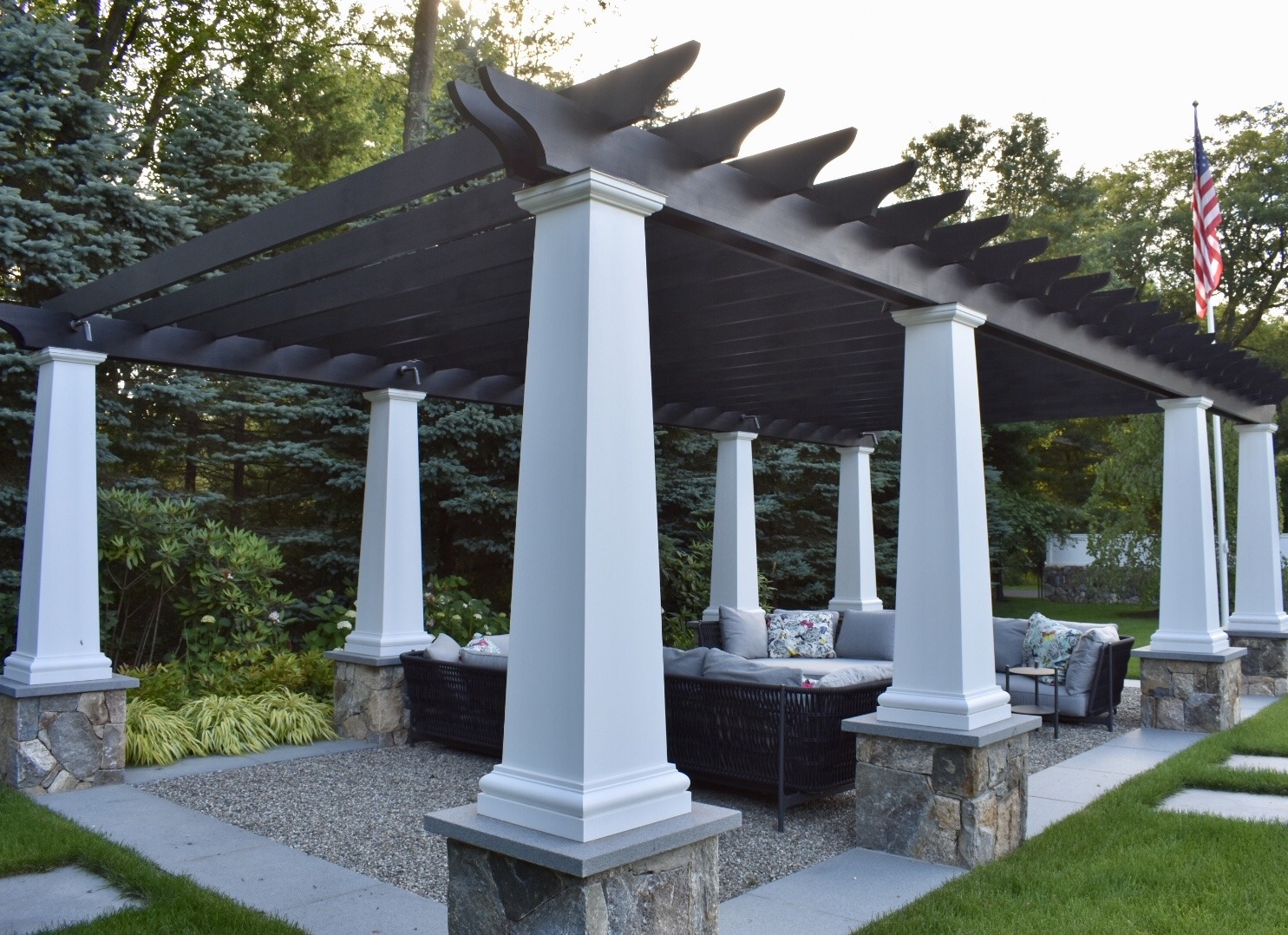An outdoor pergola with white columns and black roof slats over a seating area, surrounded by landscaped greenery and an American flag visible.