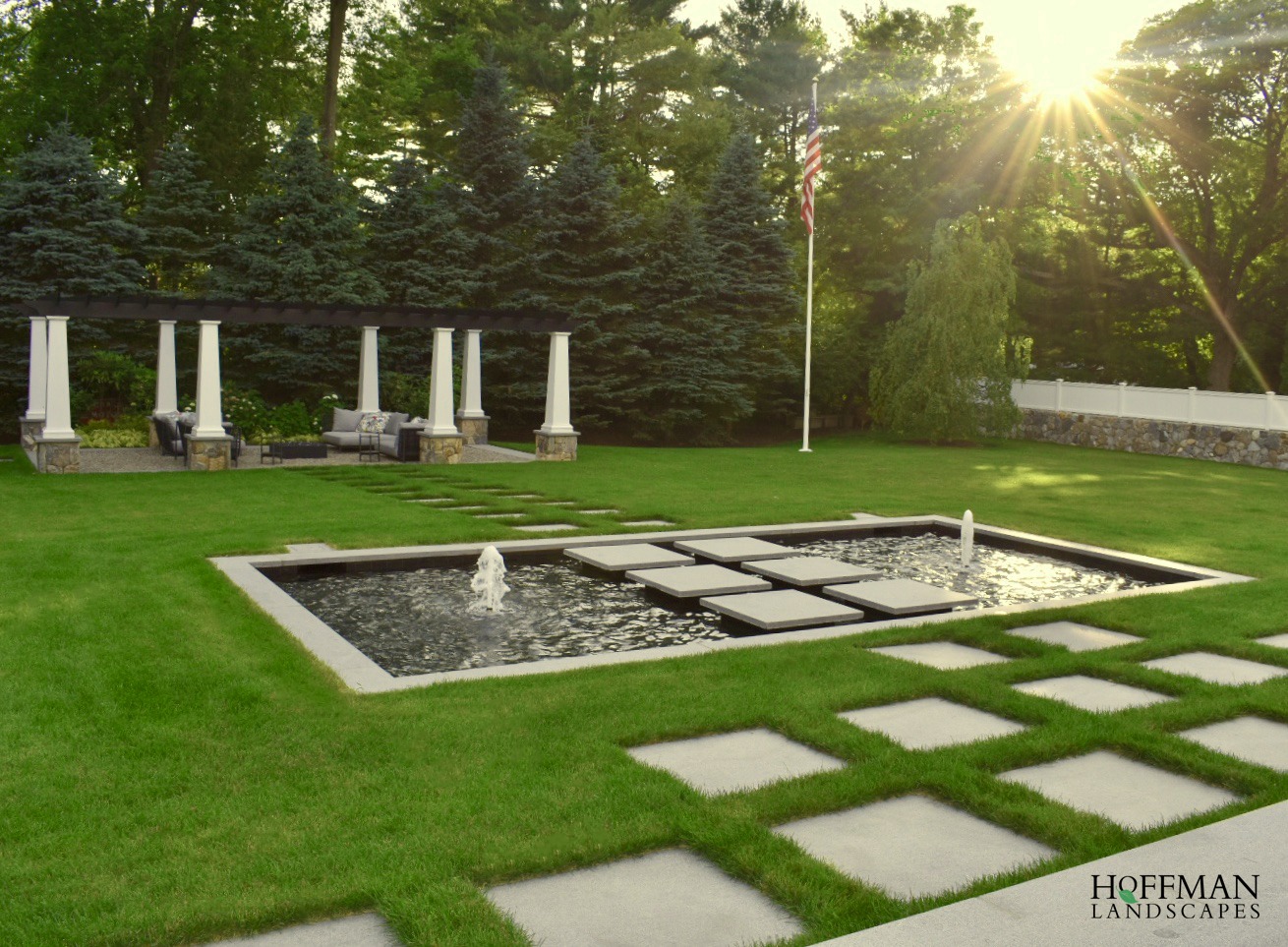 An elegant garden with a water feature and stepping stones, framed by tall columns, plush greenery, and a flagpole under a radiant sun.