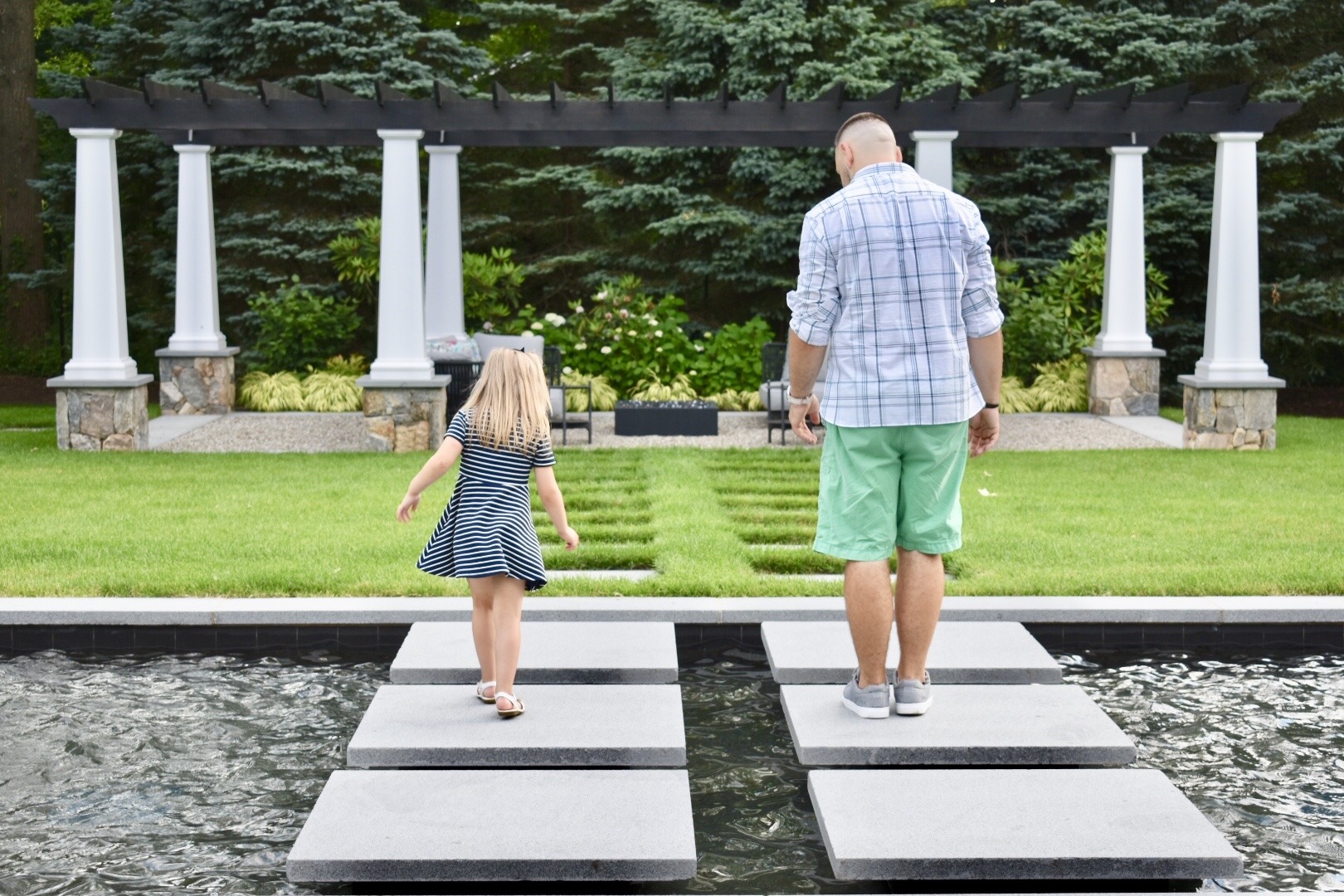 A person and a young child walk on stepping stones across water in a landscaped garden with white pillars and lush greenery in the background.
