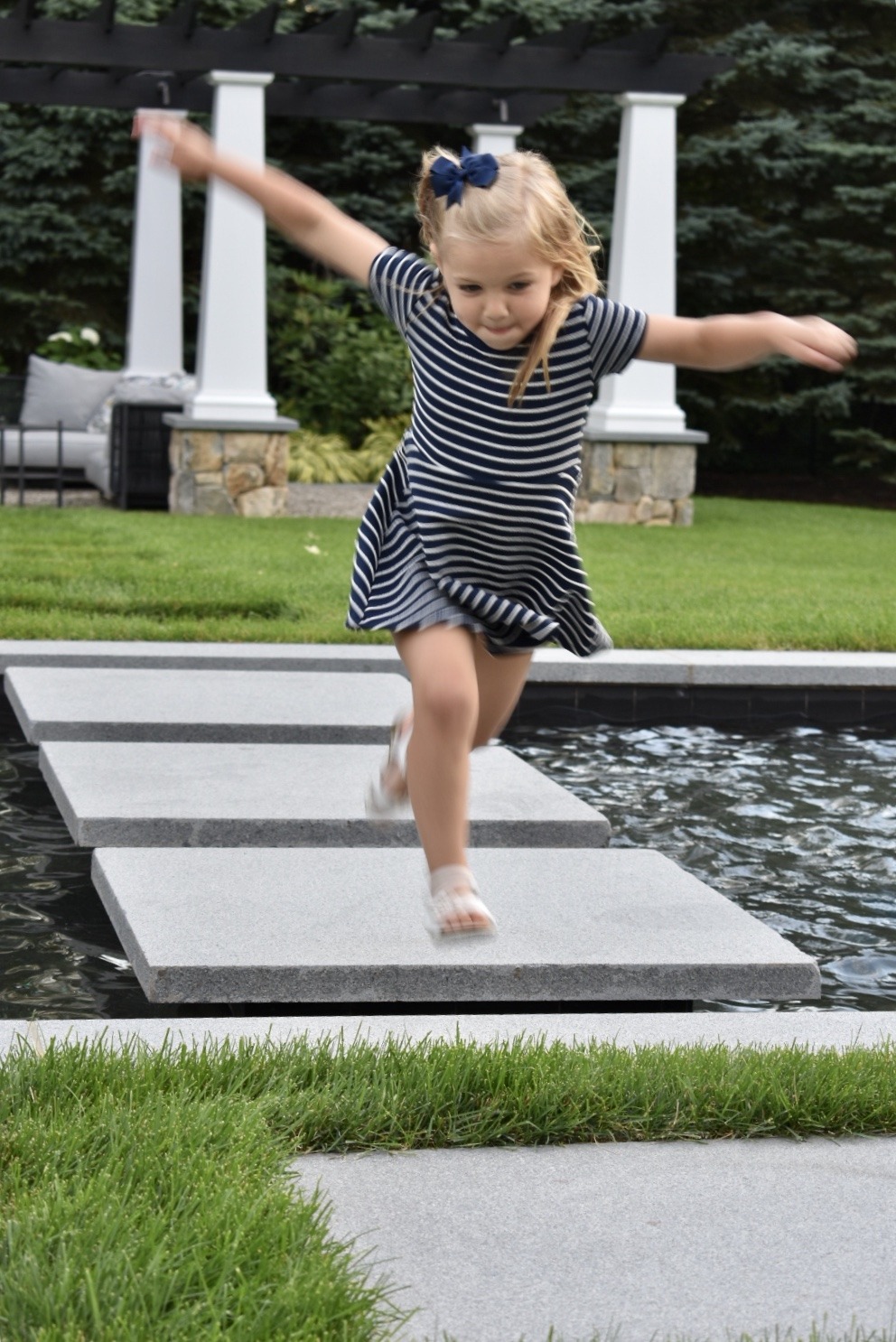 A young child joyfully balances on stepping stones across a water feature in a garden, wearing a striped dress, with trees and a pergola backdrop.